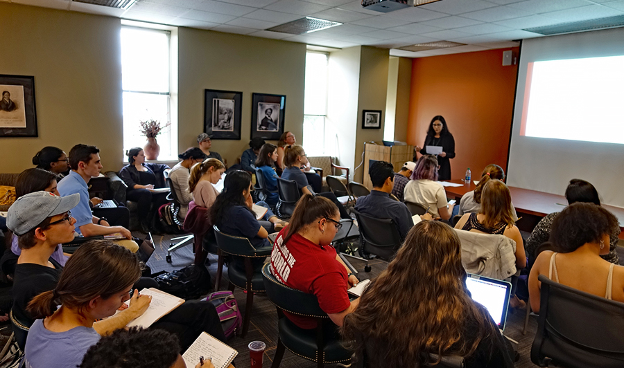 The backs of approximately two dozen students seated facing Dr. Paula Moya, who is standing and reading from a paper at the front of a large conference room.