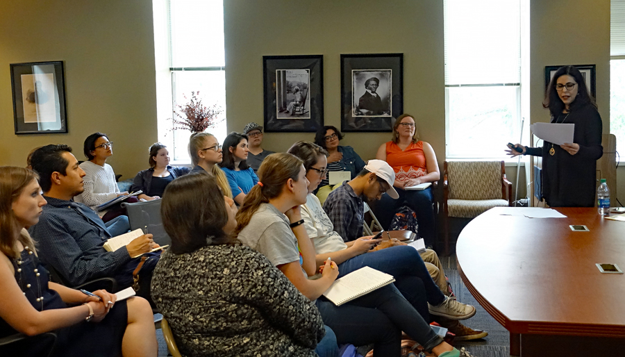 Approximately a dozen students seated in rows around a large conference room, listening attentively to Dr. Paula Moya as she reads from a paper.