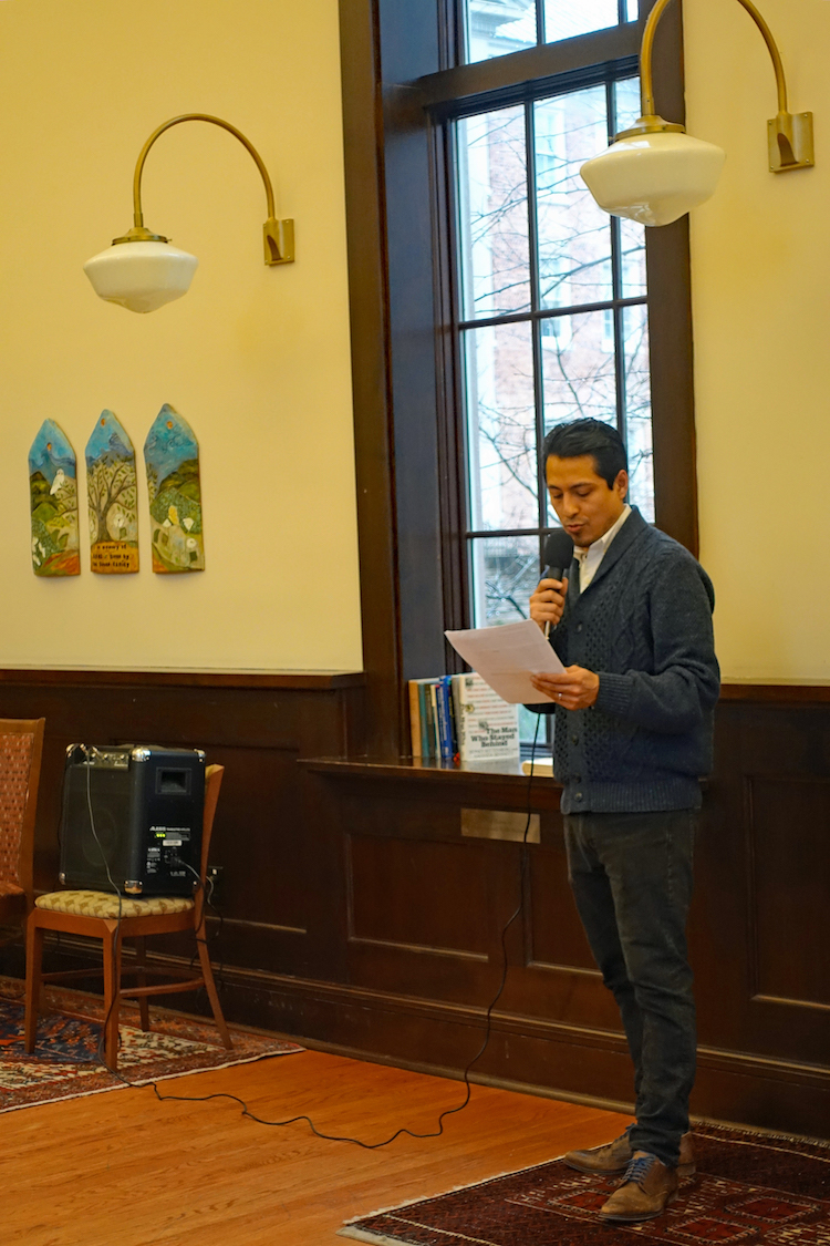 Latina/o Studies Program graduate assistant Geovani Ramírez reading from a paper and speaking into a microphone as he stands at the front a large room.