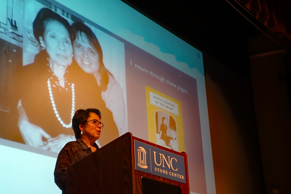 Cherríe Moraga speaking a podium before a projected presentation slide that contains photos of Moraga with her mother and the words, “I return through these pages.”